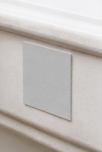 A square, light gray textured tile or panel mounted on a beige stone or concrete surface with decorative molding.