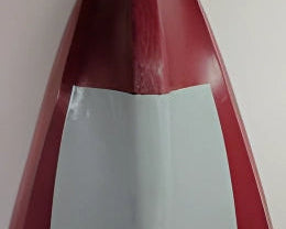 A close-up of a red surfboard or similar longboard with a large white rectangular patch on the underside, photographed against a plain light background.