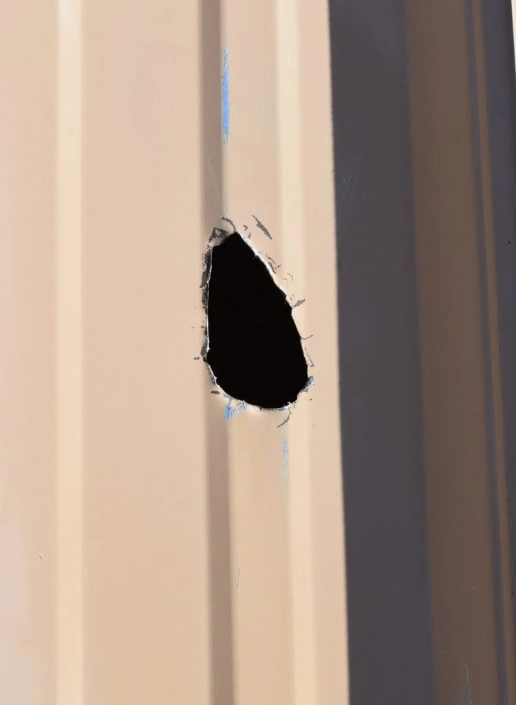 A close-up of a beige or cream-colored metal siding panel with a simulated bullet hole effect in the center.