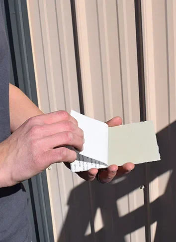 A person's hands holding an open, UV repair patch showing a light sage green color, set against a background of beige vertical siding panels with strong sunlight and shadows.