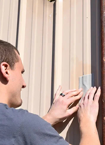 A man in a blue-gray t-shirt is shown from the side, applying a piece of gray tape to a vertical beige corrugated metal wall panel.